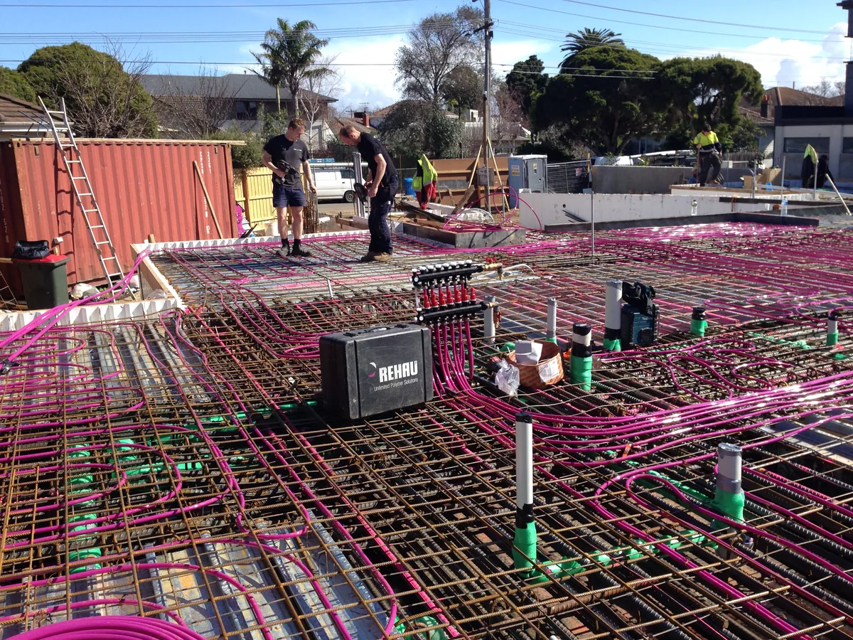 Workers beside pink pipe loops, a manifold and pipe risers laid across steel slab reinforcement