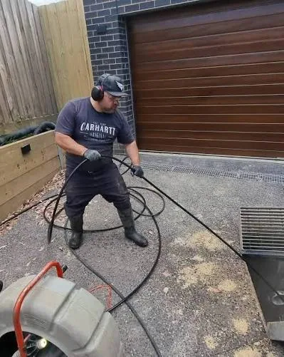 Worker handling a drain-cleaning hose beside a driveway drainage grate